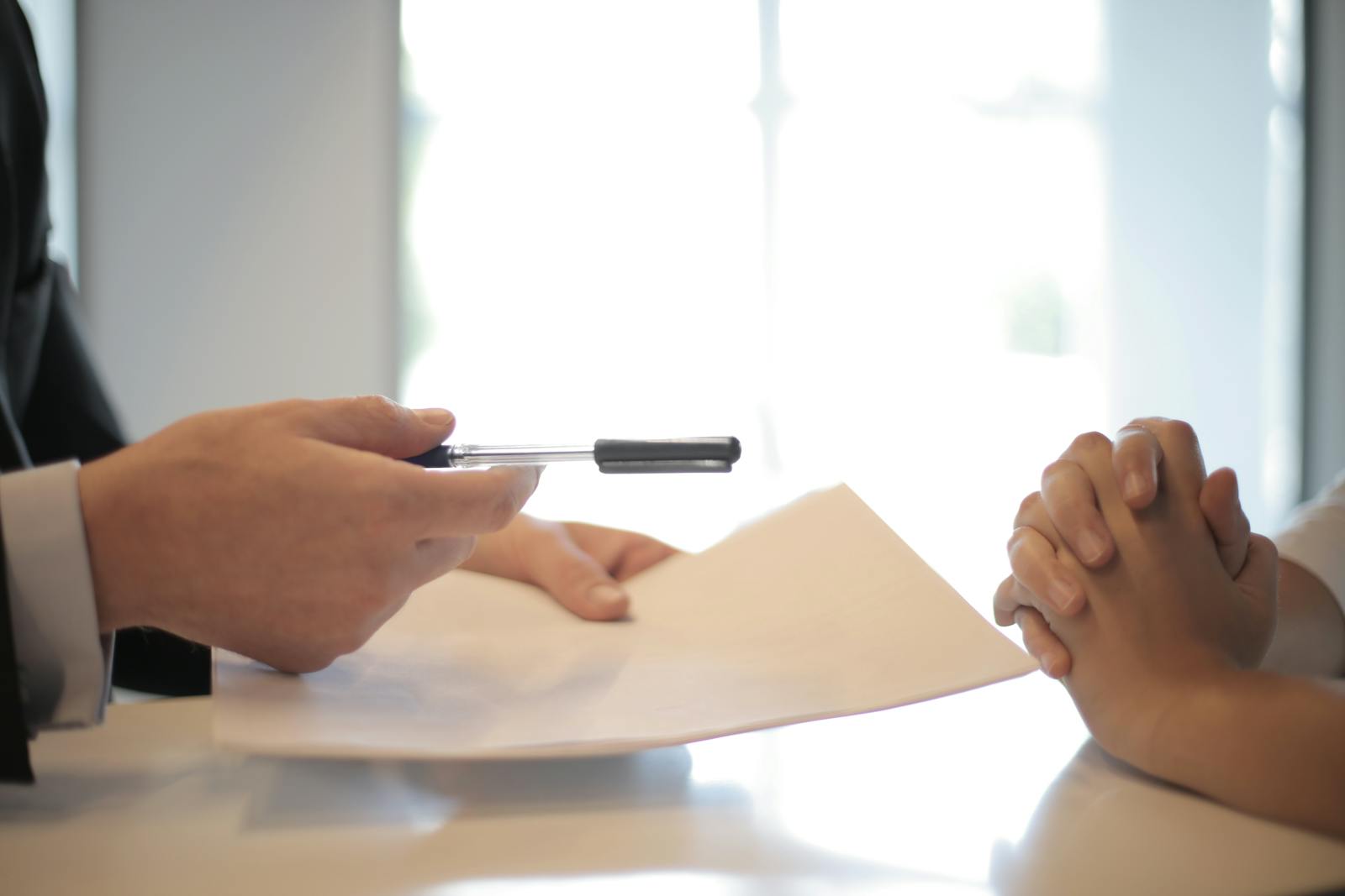 One person is handing over a document to another individual, who is sitting with their hands clasped together, possibly in an attentive or formal posture.